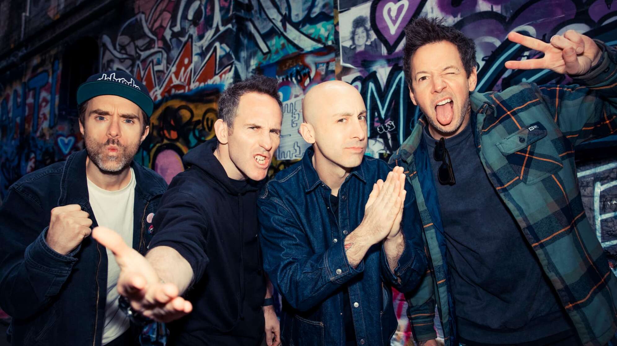 die vier Bandmitglieder von Simple Plan stehen vor einer Wand mit Graffiti und blicken in die Kamera