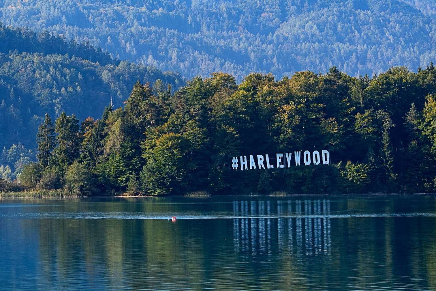 Großes #HARLEYWOOD Schild im Wald oberhalb des Faaker Sees in Kärnten, Österreich, spiegelt sich im Wasser – idyllische Kulisse der European Bike Week von Harley-Davidson.