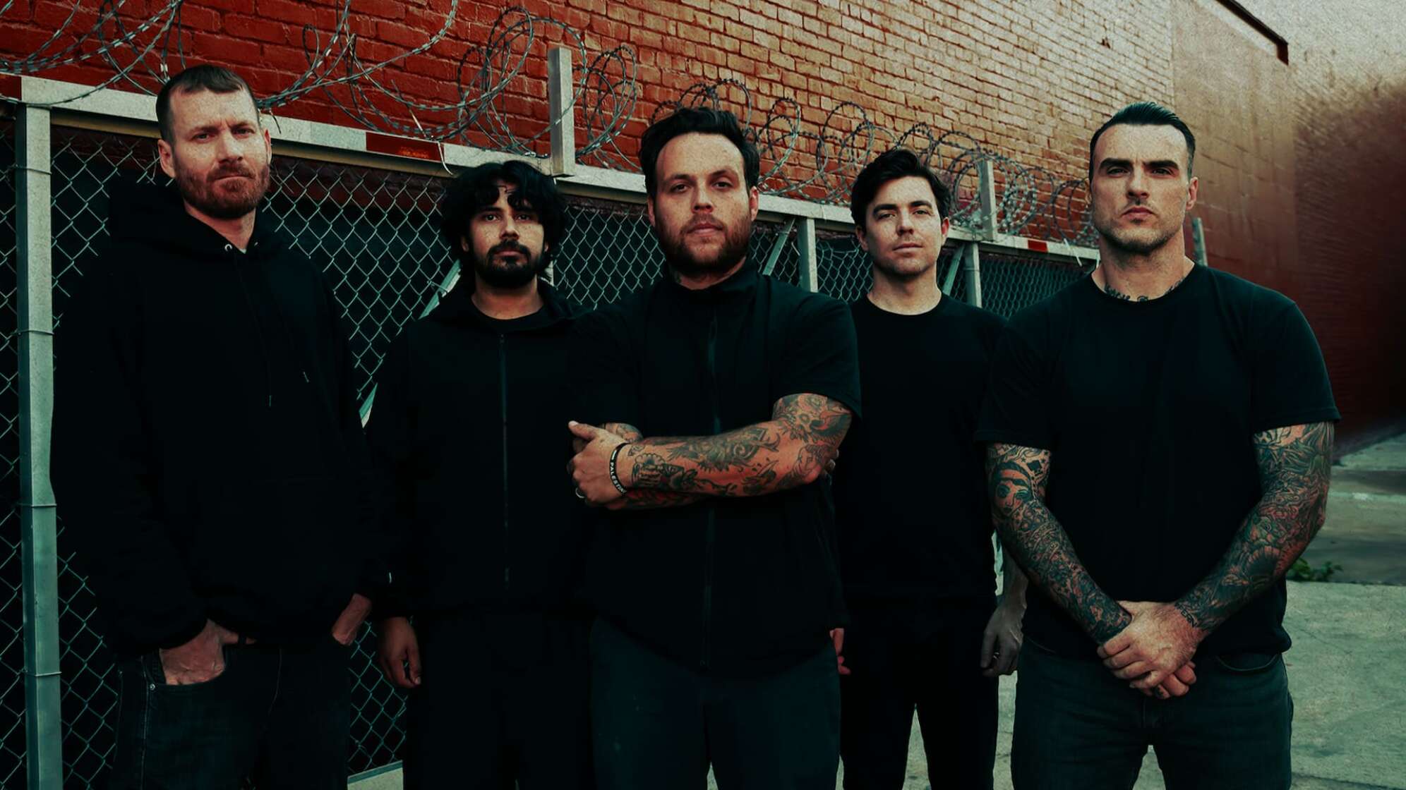 die Bandmitglieder von Stick To Your Guns vor einer Ziegelwand mit Maschendrahtzaun