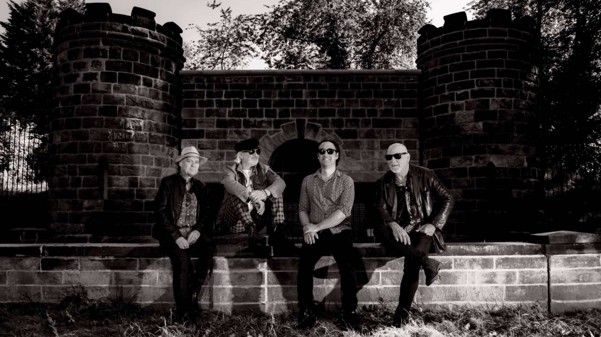 die vier Bandmitglieder von The Mission sitzen auf einer Ziegelwand, Foto in Schwarz-Weiß