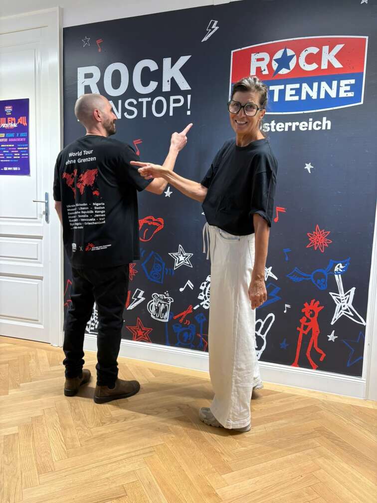 Andi Kaspar und Doris Burtscher zeigen auf das ROCK ANTENNE Österreich Logo und die Rock ohne Grenzen T-Shirts
