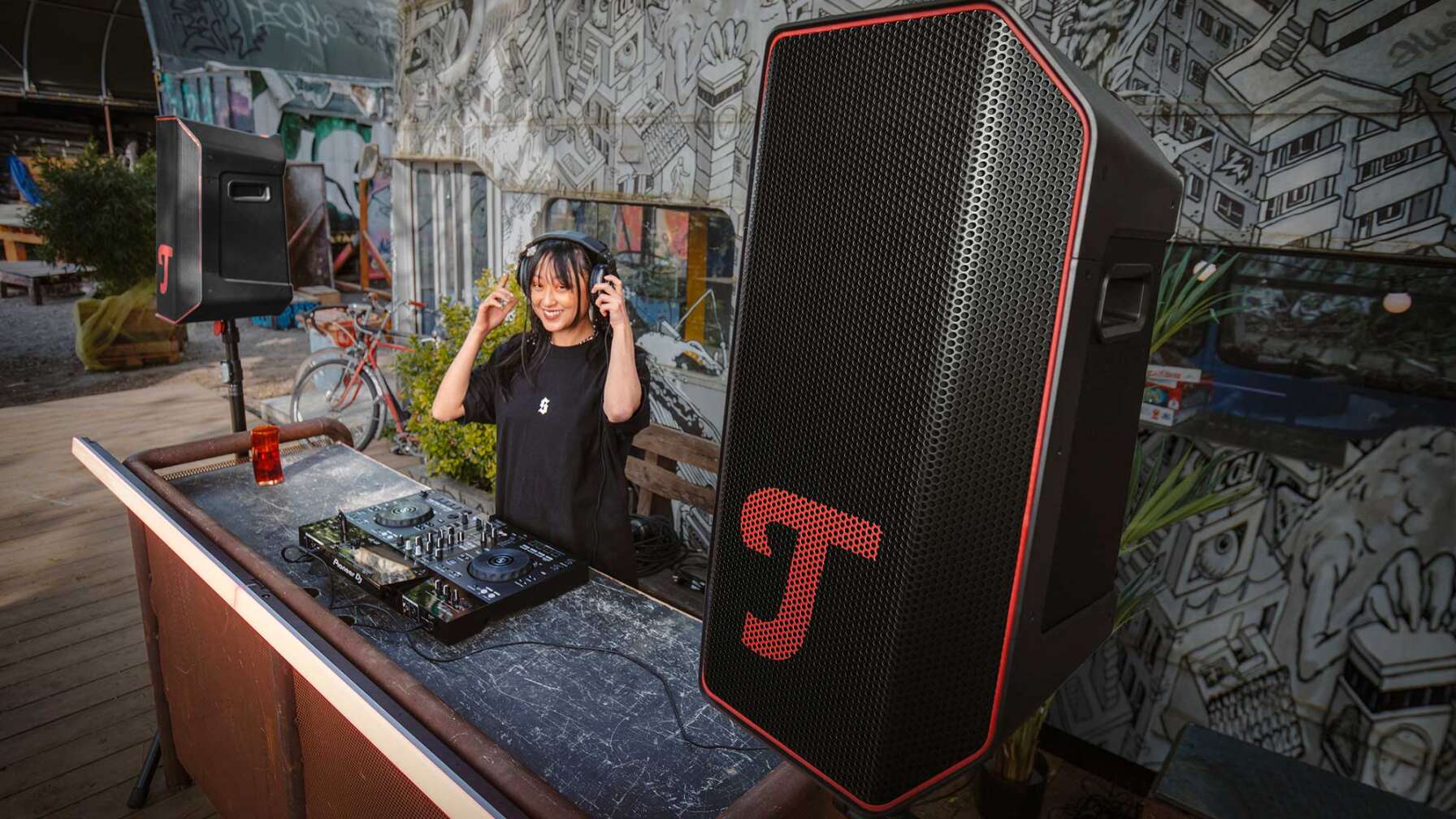 Eine junge Frau mit Kopfhörern steht an einem DJ-Pult, das an einen Lautsprecher der Marke Teufel angeschlossen ist.