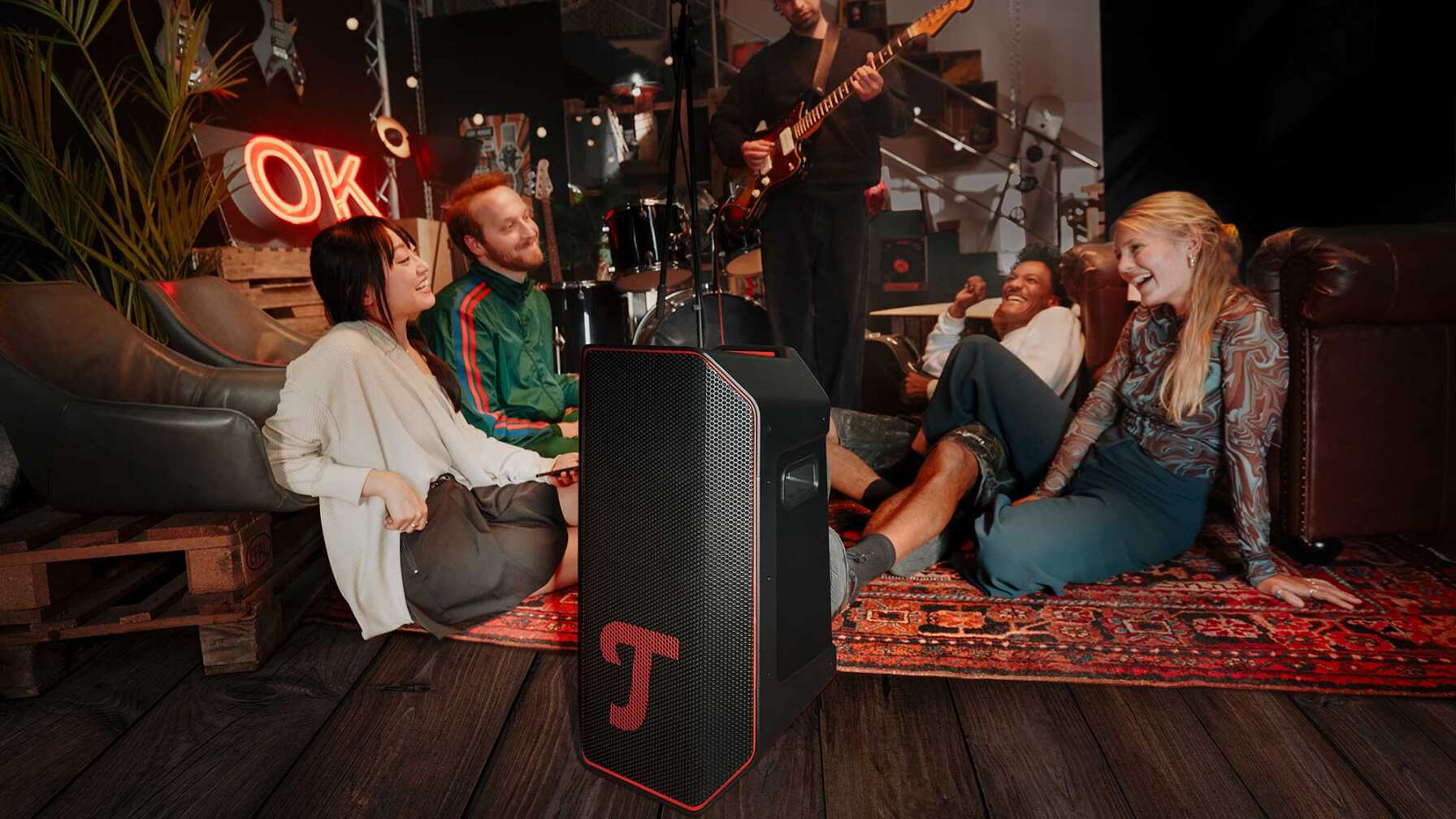 Eine Gruppe junger Leute sitzen in einem alternativ eingerichteten Raum vor einem Lautsprecher der Marke Teufel. Im Hintergrund spielt ein junger Mann E-Gitarre.