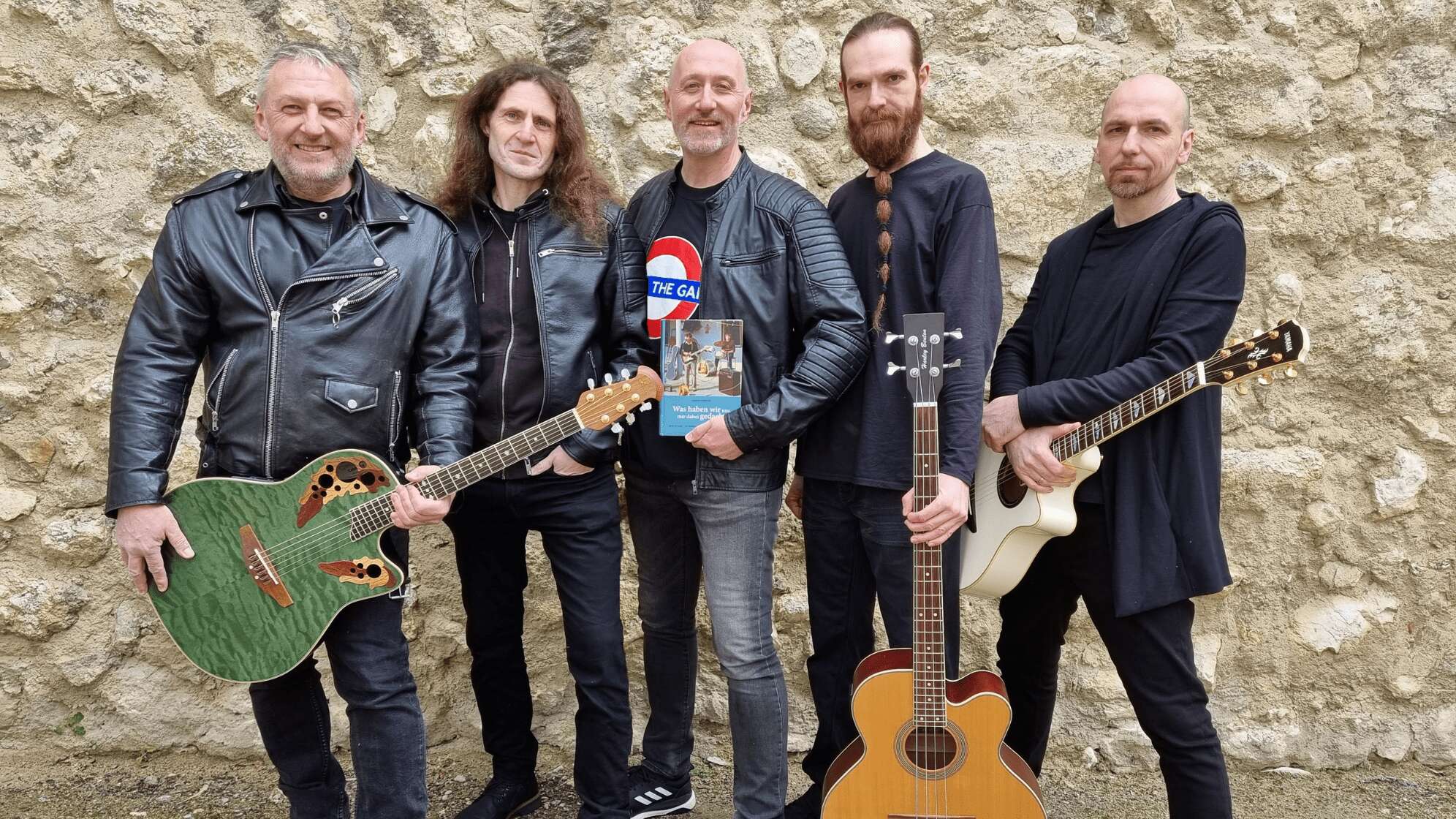 5 Männer mit Gitarren in der Hand vor einer Steinmauer.