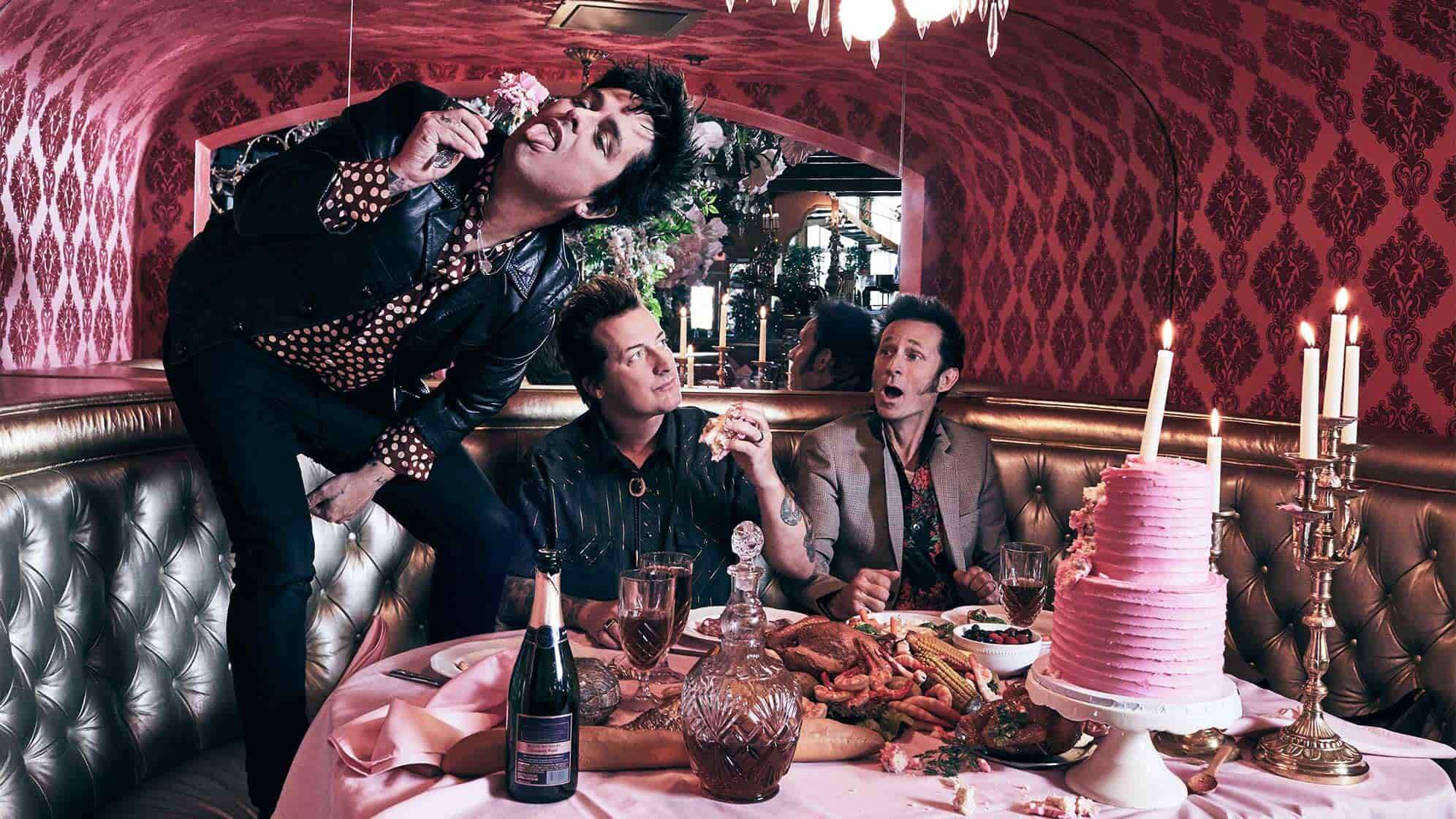 Ein Bandfoto der Band Green Day aus dem Jahr 2020, an einem überladenen Esstisch mit Grimassen