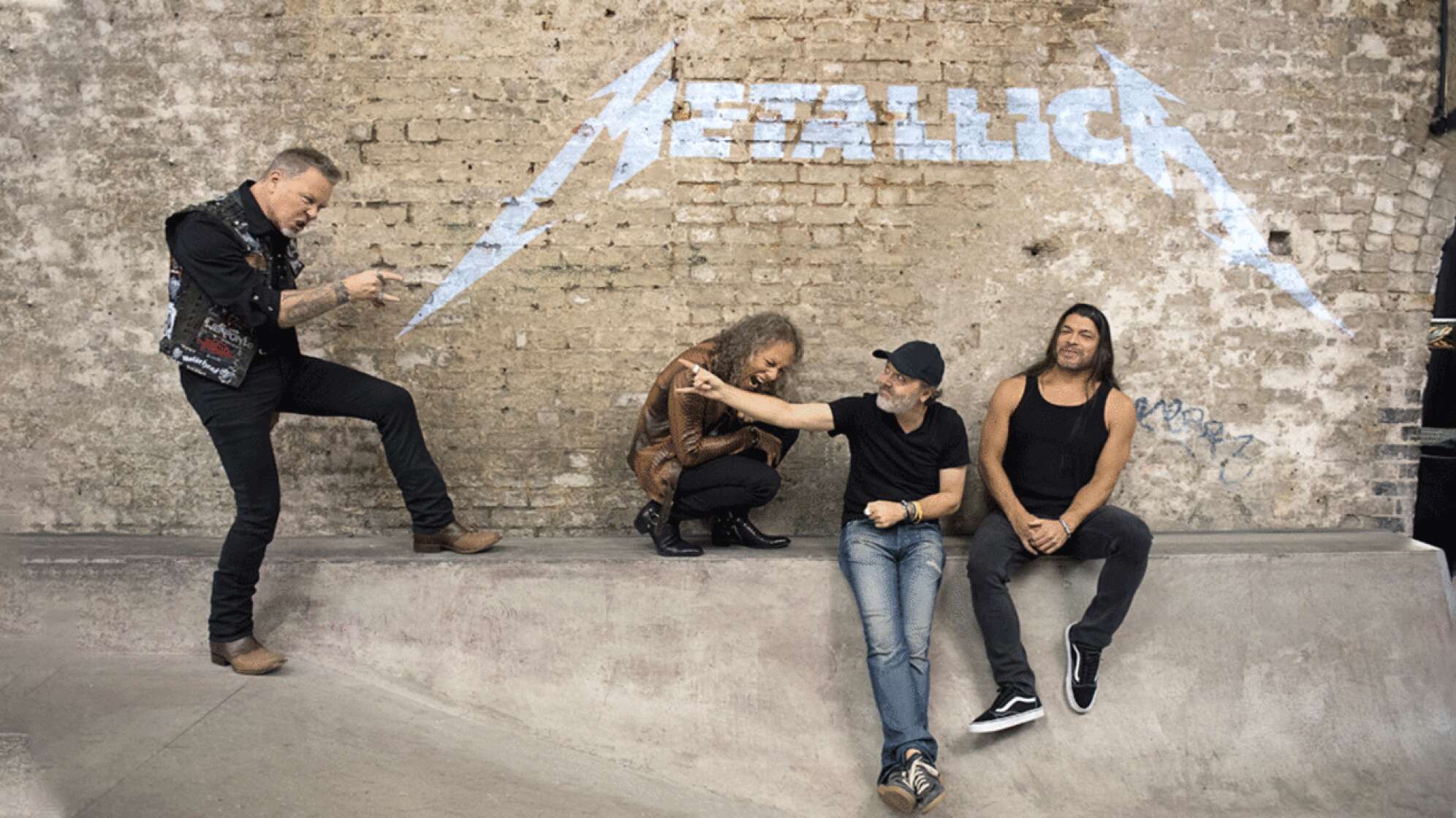 Metallica posieren vor einer Backstein mit ihrem Logo